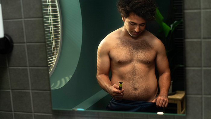 Topless man holds the Philips OneBlade electric razor with guard attachment before trimming the hair below his waist.